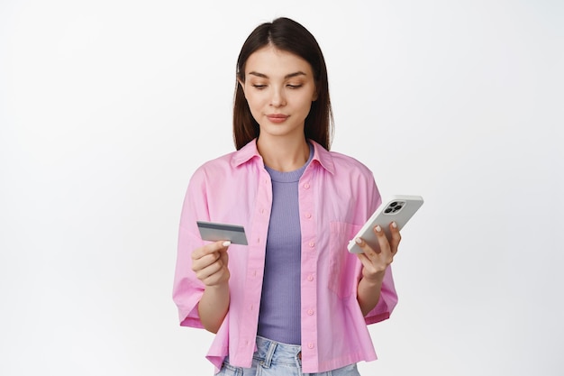 Image of smiling woman making purchase on mobile phone looking at credit card while order something on smartphone app standing over white background
