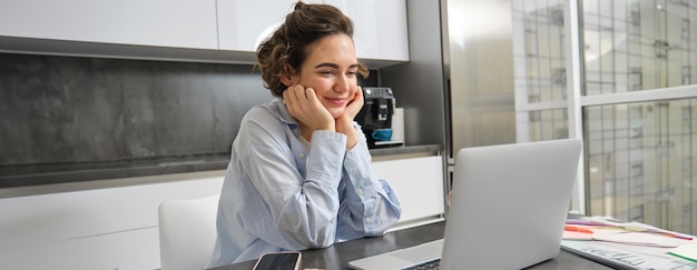 Free photo image of smiling woman studying on remote looking at laptop while sitting at home watching webinar