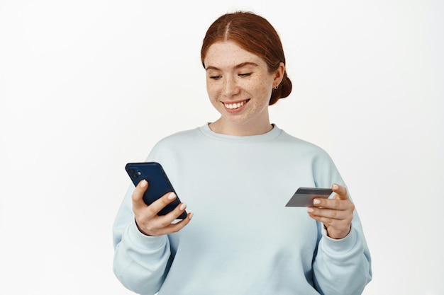 Image of smiling young redhead woman register her credit card in application, looking at mobile phone screen, holding discount card, standing against white background.