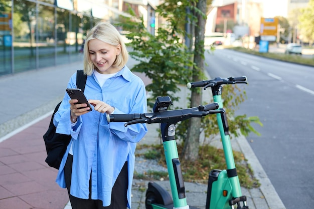 Image of smiling young woman heading to work renting using mobile phone app to unlock street scooter