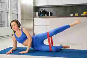 Free photo image of sporty girl doing aerobics using resistance band on legs workout at home listens to music