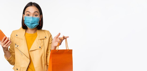 Изображение стильной азиатской девушки-покупателя, держащей смартфон и сумку для покупок без логотипа магазина в медицинской маске на белом фоне covid19