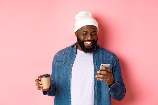 テイクアウトのコーヒーを飲み、電話でメッセージを読んで、ピンクの背景の上に立って笑っているスタイリッシュな黒人男性のヒップスターの画像。