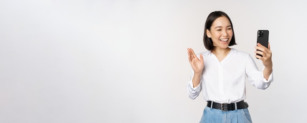 Изображение стильной современной азиатской женщины, машущей рукой перед камерой смартфона в видеочате, разговаривающей с человеком в приложении для мобильного телефона на белом фоне