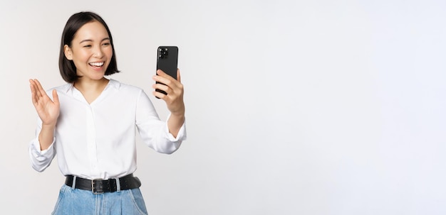 Image of stylish modern asian woman waving hand at smartphone front camera video chat chatting with person on mobile phone application white background