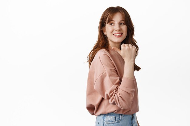 Image of stylish redhead teen girl, look behind her shoulder and smiling, pointing finger left at copy space advertisement, standing against white background