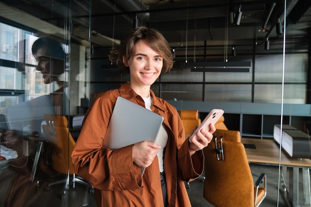 Free photo image of stylish working woman checking her messages on smartphone holding laptop standing in an