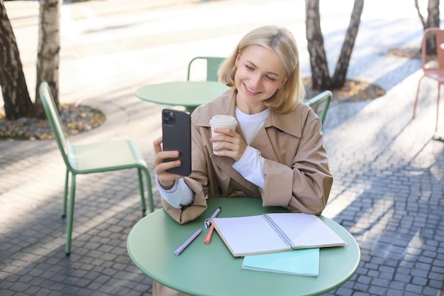Изображение стильной молодой студентки, делающей селфи в кафе на улице, позирующей со своим любимым напитком