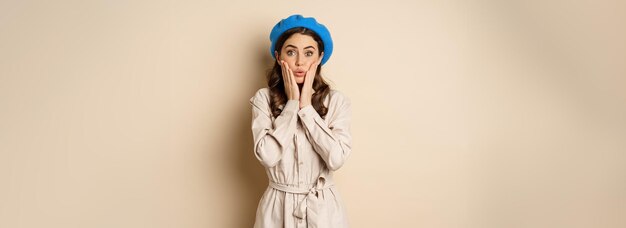 image of stylsh beautiful woman looking surprised shocked reaction at camera posing in trench coat against beige background