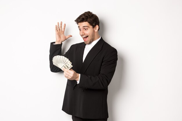 Image of successful businessman in black suit, looking at money and triumphing, earn cash, standing over white background.