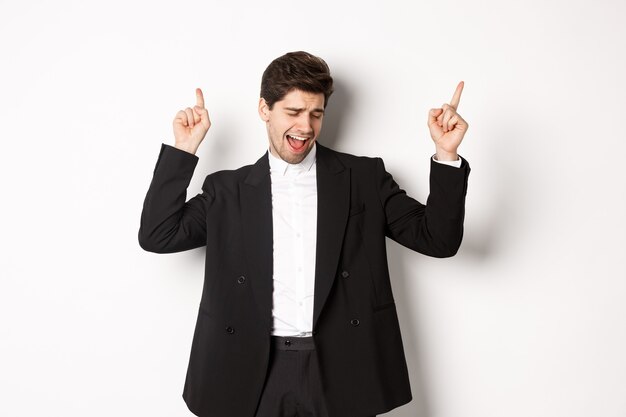 Image of successful handsome businessman dancing, enjoying party, pointing fingers up and having fun, standing over white background, wearing black suit.