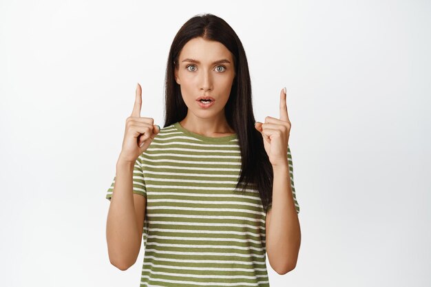 Image of surprised adult woman pointing up looking in awe at camera standing in tshirt over white background