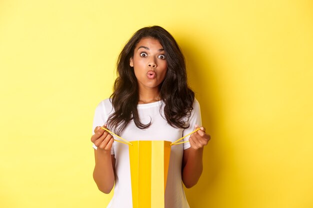 놀란 아프리카계 미국인 소녀의 이미지는 휴일에 선물을 받고 쇼핑백을 열고 놀란 표정으로 노란색 배경 위에 서 있습니다.