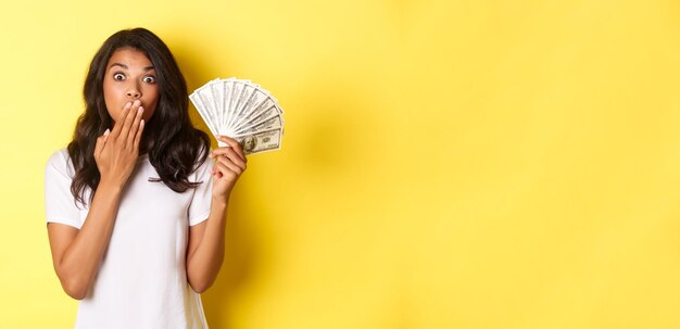 Image of surprised africanamerican girl winning money gasping amazed standing over yellow background