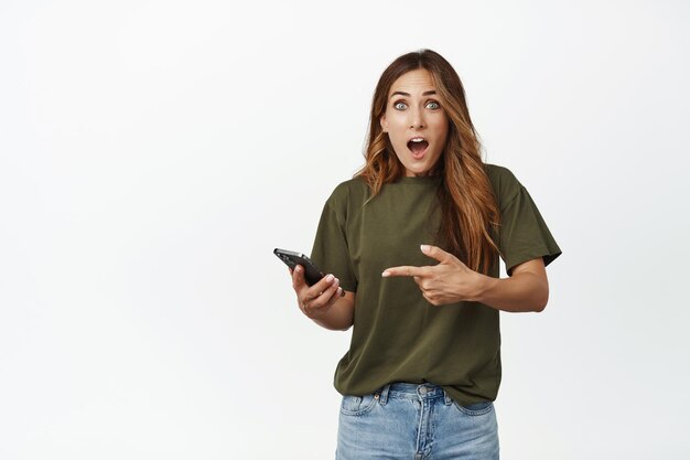 Image of surprised and amazed woman gasp fascinated, pointing finger at smartphone, using awesome app, found smth online, standing against white background