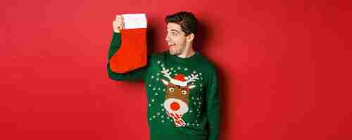 Free photo image of surprised and amused man in green sweater looking at christmas stocking with presents and s...