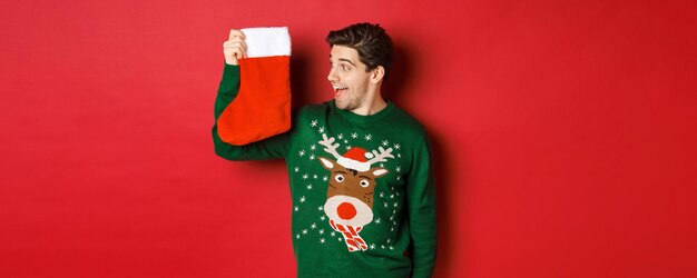 선물과 선물이 있는 크리스마스 스타킹을 보고 있는 녹색 스웨터를 입은 놀랍고 즐거운 남자의 이미지...