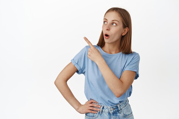 Image of surprised blond girl, gasping in awe, pointing at upper left corner, looking at price sale, discount advertisement, standing in t-shirt over white background.