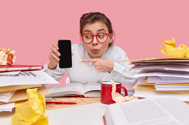 Image of surprised European woman has shocked expression, has folded lips, shows something on blank space, carries modern cell phone