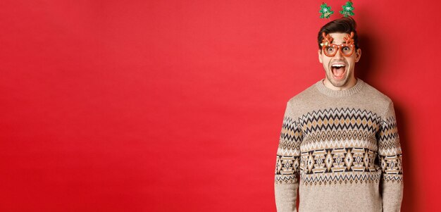 크리스마스 스웨터 드롭 턱이 있는 파티 안경을 쓰고 빨간색 배경에 서 있는 놀라운 새해 프로모션 제안을 바라보는 놀랍고 흥분된 잘 생긴 남자의 이미지