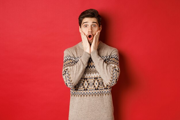 Image of surprised handsome guy reacting to cool new year promo offer, gasping amazed, wearing christmas sweater, standing over red background