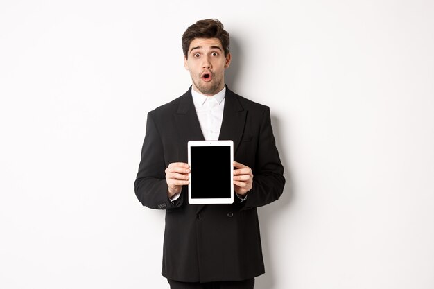 Image of surprised handsome man in black suit, showing digital tablet screen and looking amazed, standing against white background