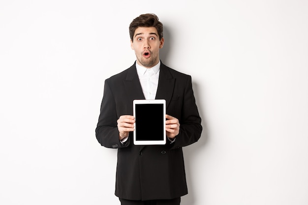 Image of surprised handsome man in black suit, showing digital tablet screen and looking amazed, standing against white background