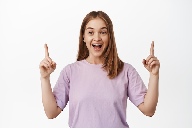 Image of surprised and happy authentic woman pointing fingers up, showing advertisement, smiling amazed, laughing happily, standing against white background. Copy space
