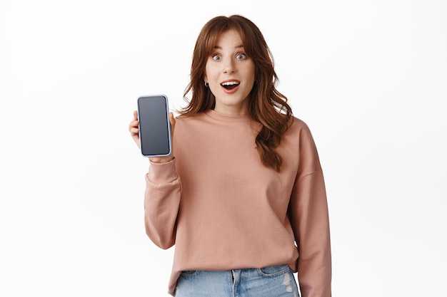 Image of surprised teenage girl drop jaw, showing advertisement on smartphone screen, recommend app on phone, standing in sweatshirt against white background