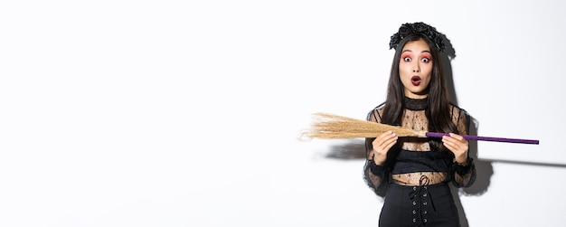 Image of surprised woman in halloween costume holding witch broom and looking at camera amazed