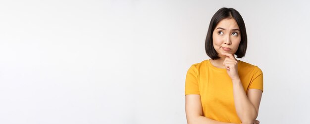 Изображение думающей азиатской женщины, смотрящей в сторону и обдумывающей принятие решения, стоящей в желтой футболке