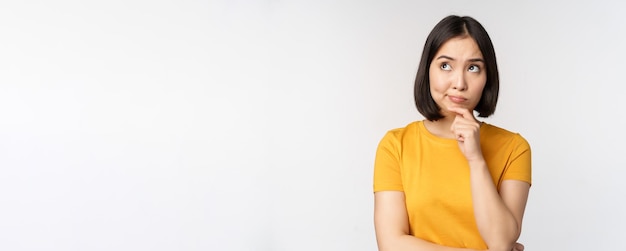 Image of thinking asian woman looking aside and pondering making decision standing in yellow tshirt