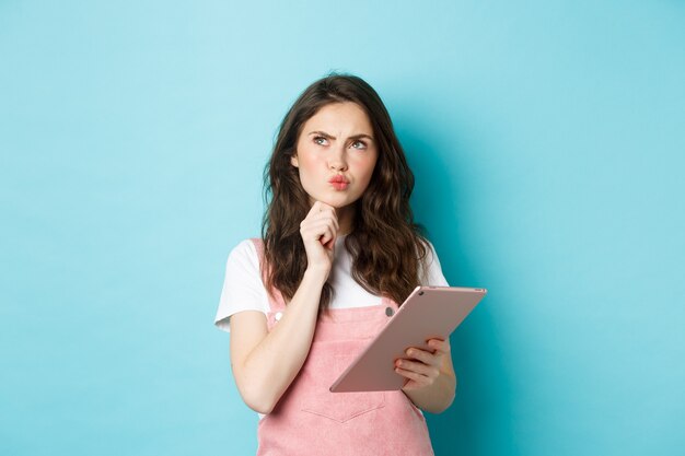 Image of thoughtful female student looking at upper left corner and thinking, holding digital tablet, making hard choice, standing over blue background.