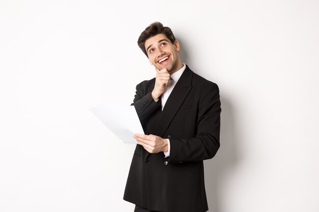 Image of thoughtful, handsome businessman in black suit, holding document and looking at upper right corner, thinking and smiling, standing against white background