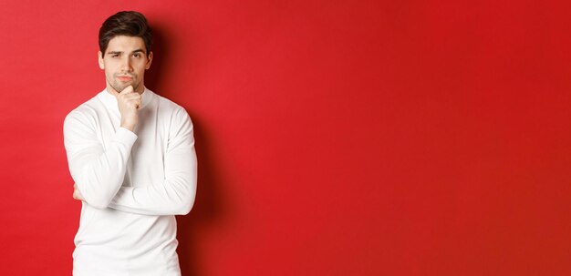 Image of thoughtful handsome man making assumption, thinking and looking at camera, standing in white sweater against red background.