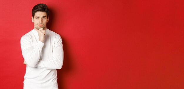 Изображение вдумчивого красивого мужчины, делающего предположения, думающего и смотрящего в камеру, стоящего в белом свитере на красном фоне.