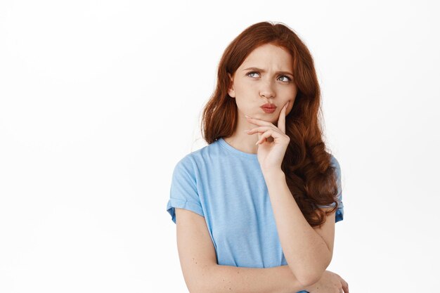 Image of thoughtful hesitant girl with red hair making decision, frowning perplexed and thinking, touching lip pondering, standing against white background.