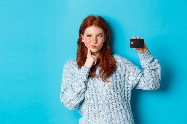Изображение вдумчивой рыжей девушки, думая о покупках, показывая кредитную карту и размышляя, стоя на синем фоне.