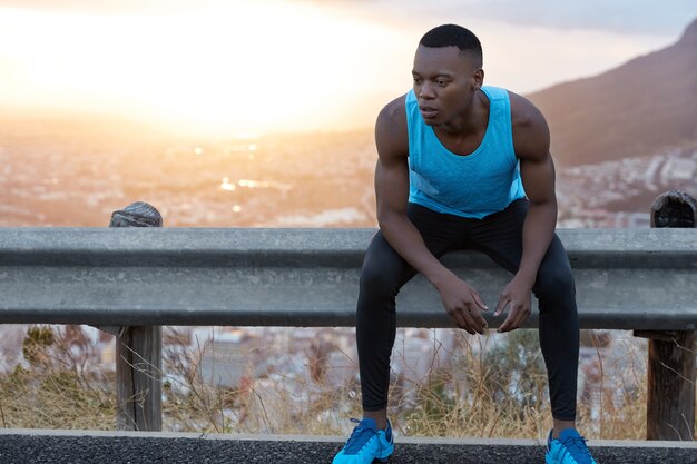 Image of tired African American man with thoughtful expression, keeps gaze down, feels tired after intense training, sits at road sign, beautiful sunrise  with copy space for information