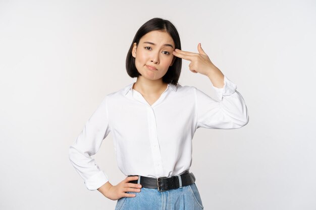 Image of tired asian woman annoyed by smth pointing fingers at her head standing over white background Copy space