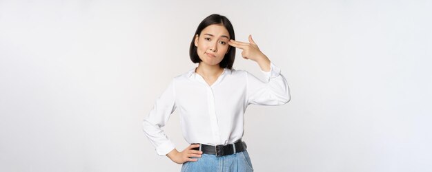 Image of tired asian woman annoyed by smth pointing fingers at her head standing over white background Copy space