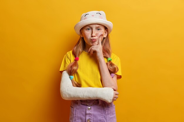 Image of toughtful little girl keeps finger on cheek and thinks deeply, has displeased grimace, contemplates how to recover quickly, has broken arm during playing on playground and falling from swing