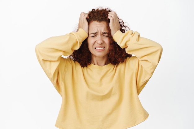 Immagine di una donna rossa turbata e frustrata che tiene le mani sulla testa e fa una smorfia in preda alla disperazione, angosciata, brutta notizia, in piedi ansiosa su sfondo bianco