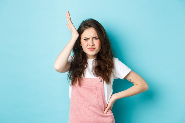 Image of troubled young girlfriend holding hand on head and frowning, remember or forget something important, standing bothered against blue background.