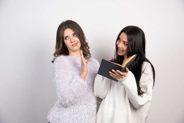 Image of two best friends standing together and posing with book on white wall .