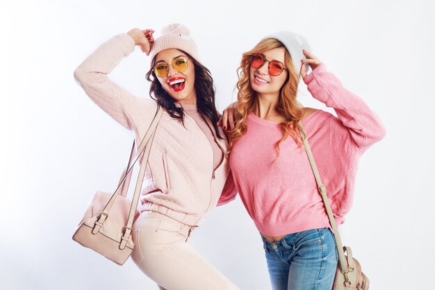 Image of two girls, happy friends in stylish pink clothes and hat spelling funny  the together. White background. Trendy hat and glasses.Showing peace.