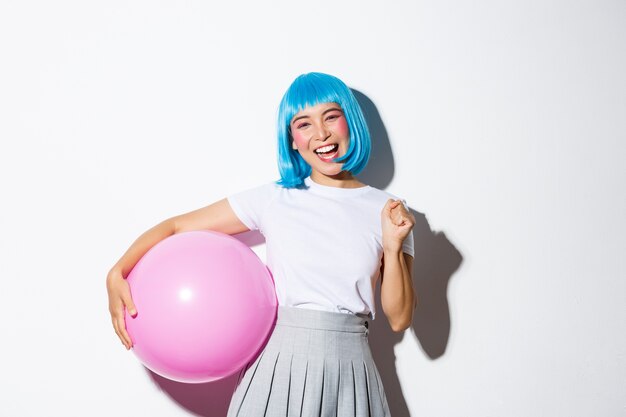 Image of winning cheerful asian girl, looking happy and triumphing, celebrating holiday, wearing party outfit and blue wig