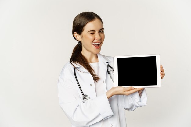 Image of woman doctor, female healthcare worker showing online medical website, digital tablet screen and smiling, standing in white coat over white background.