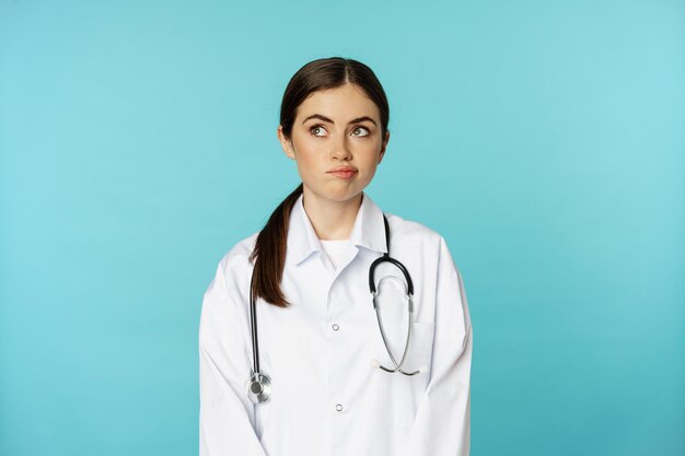 女性医師、白い白衣を着た女性の内側のスタッフ、思慮深く目をそらし、決定を下し、smthを考え、青い背景の上に立っている画像