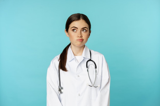 Image of woman doctor, female medial staff in white lab coat, looking away thoughful, making decision, thinking of smth, standing over blue background